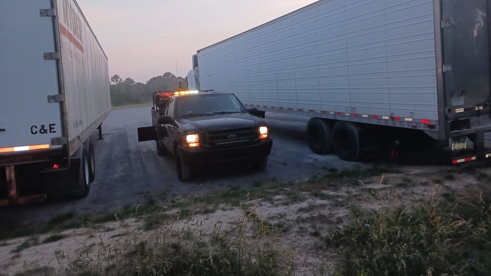 A Farmerboys service truck with its lights on, parked beside two tractor-trailers on a road call at dusk